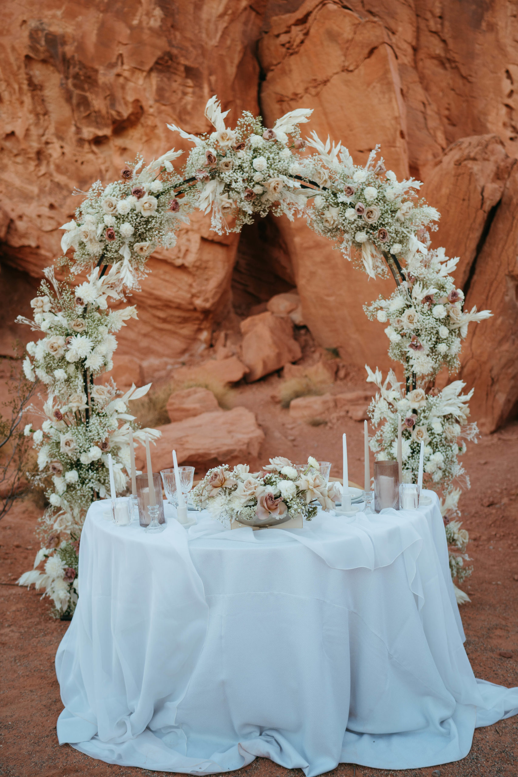 Modern Monochromatic Desert Micro-Wedding. Sweetheart table draped in white linens with monochromatic candles and votives in front of the floral covered arch in the middle of valley of fire