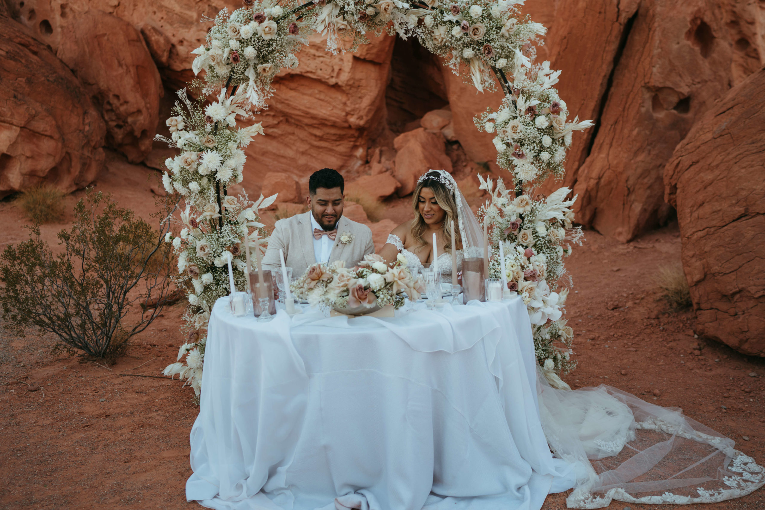 Modern Monochromatic Desert Micro-Wedding. The bride and groom at the sweetheart table draped in white linen with monochromatic candles and votives in front of the floral arch in the desert 