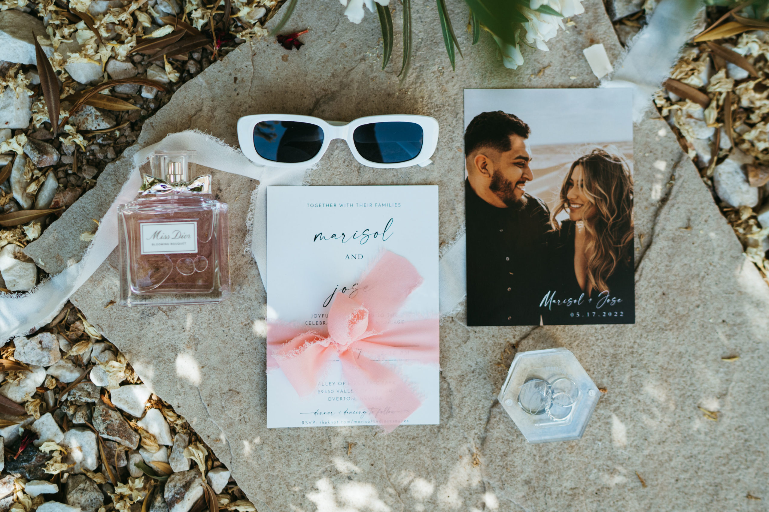 Wedding day detail photo of the couples save the day and invitation with the brides perfume of the day and sunglasses with both the bride and grooms wedding bands in a plush ring box