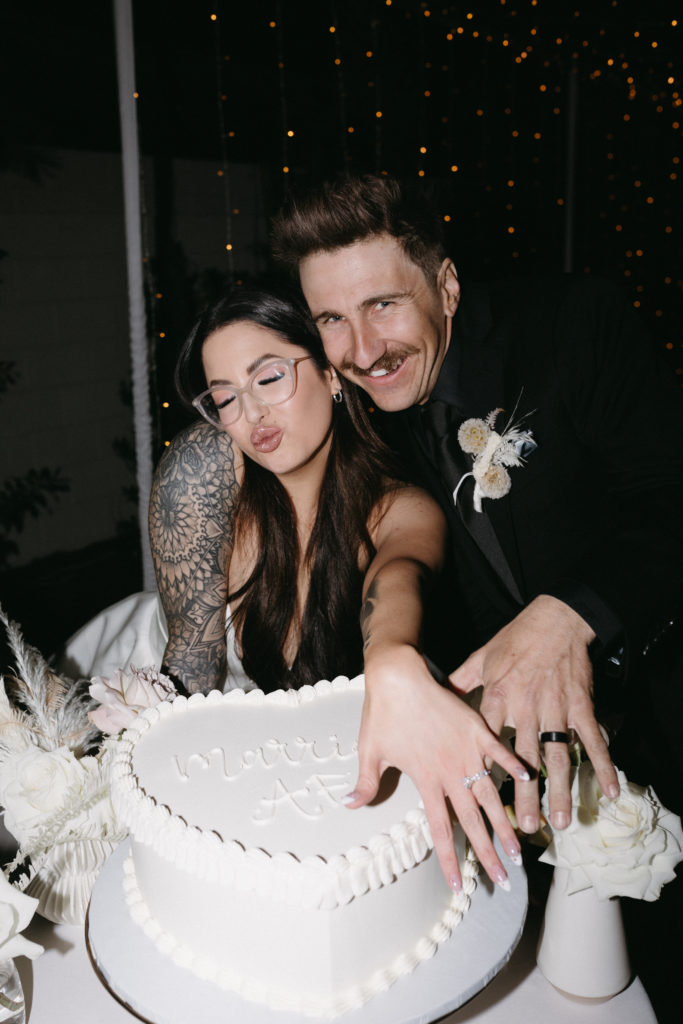 The Official List of 2023 Wedding Trends! Newlyweds standing over their wedding cake showing off their wedding bands with some night time flash photograhy 