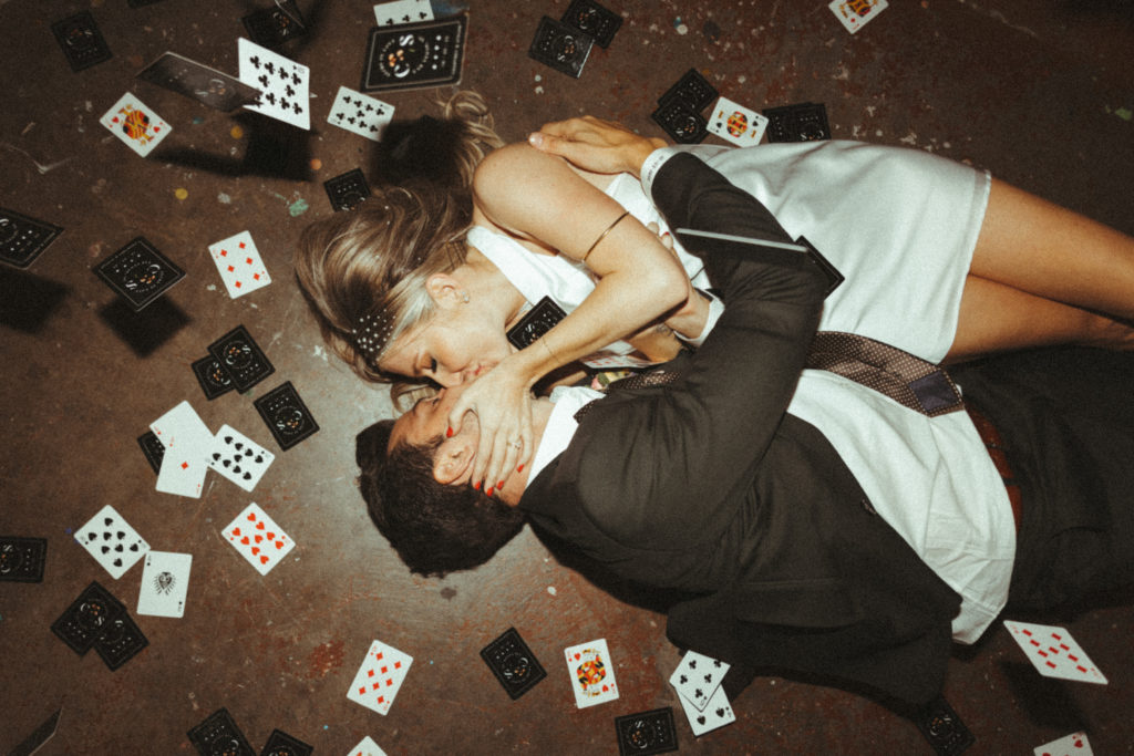 The Official List of 2023 Wedding Trends! Newlyweds lying on the ground kissing while the guest throw custom playing cards with flash photography