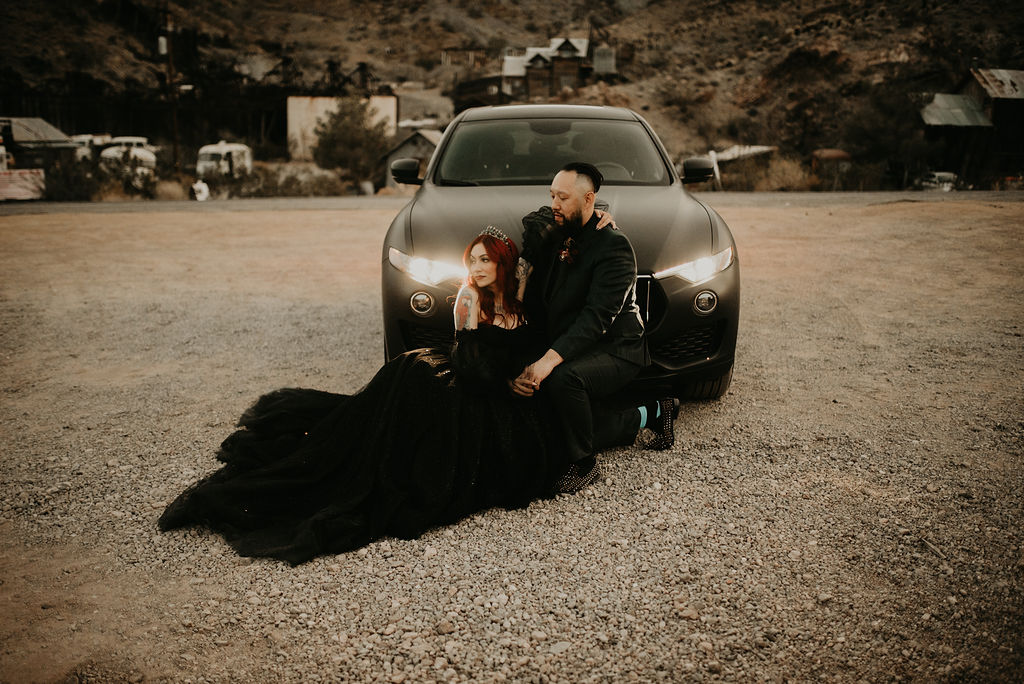 Bride laying on ground next to groom and their car 