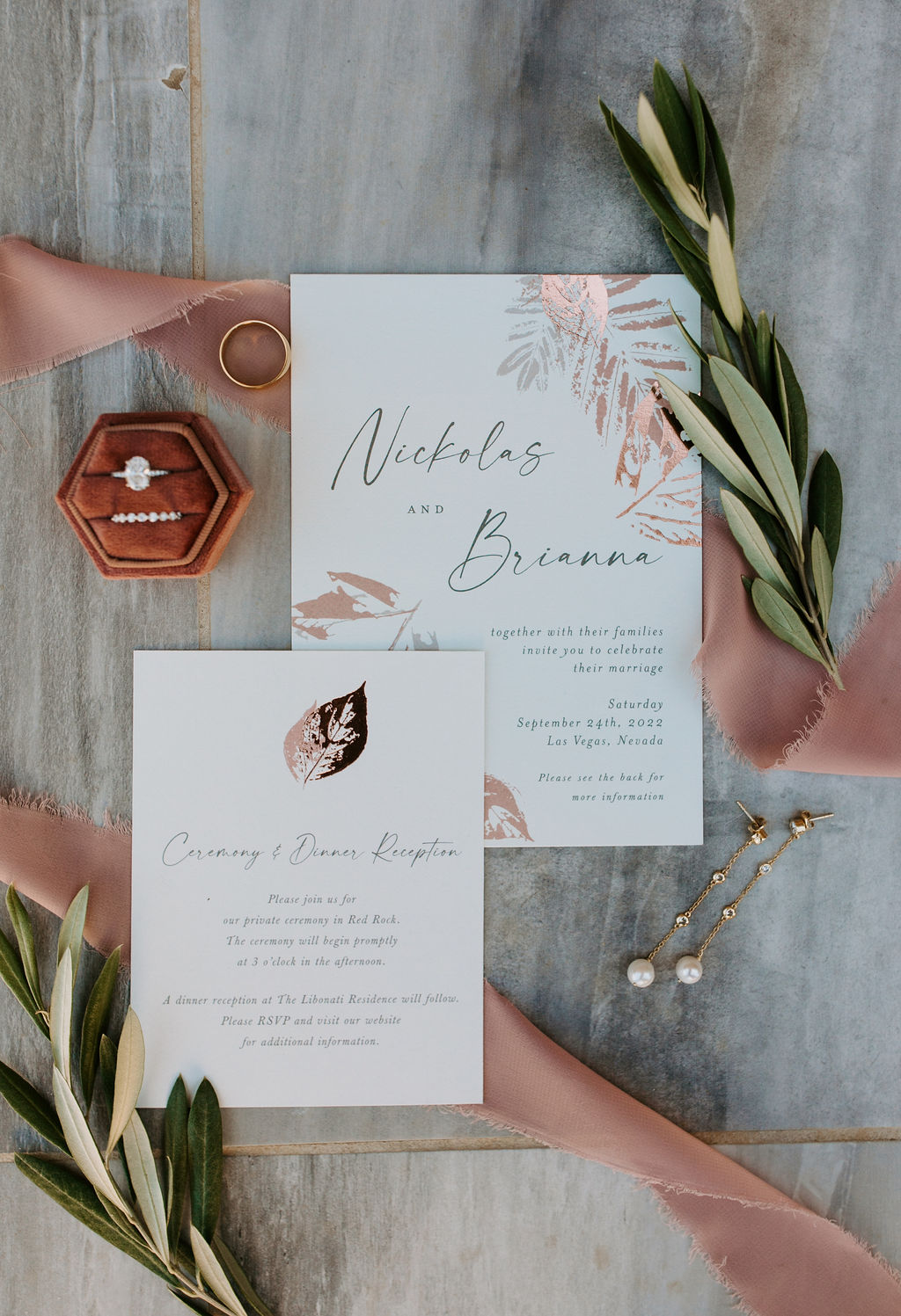 Flat lay wedding details with ring box, pink ribbon, greenery, and wedding invitations for a soft pink, mauve and sage color scheme. 