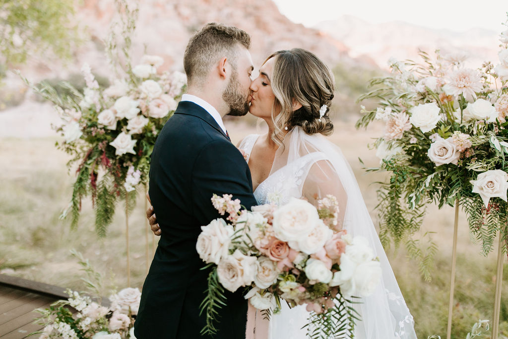 Bride and Groom kissing in front of Whimsical Ceremony Decor with Harlow Stands and Soft Pink and Sage Floral for Romantic Desert & Backyard Micro-Wedding 
