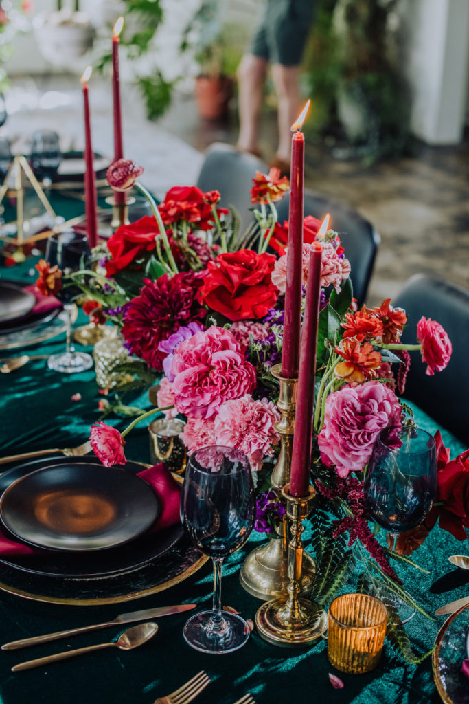 Magenta jewel toned inspired tablescape. Deep green linen paired with black plating and a silk magenta napkin and blue wine glasses. Amongst magenta taper candles and a jewel tone floral cetnerpiece. 
