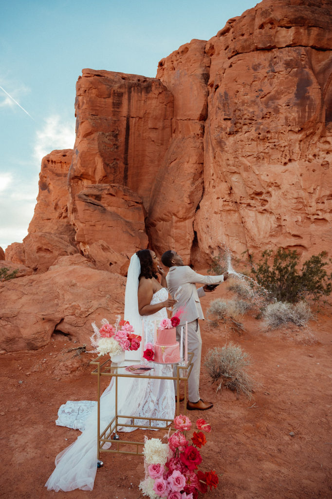 Bold & Colorful in the Vegas Desert. Newlyweds pop champagne as they stand behind the cake table 