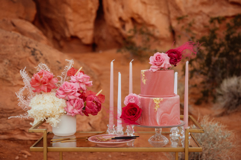 Bold & Colorful in the Vegas Desert. Gold trimmed cake table amongst the red rocks in valley of fire topped with a monochromatic floral center pieces, light pink and white votives and a two tier marbled magenta wedding cake with 24k gold flakes and florals 
