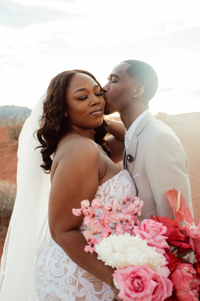 Bold & Colorful in the Vegas Desert. The groom kisses the brides cheek softly as the sun sets 
