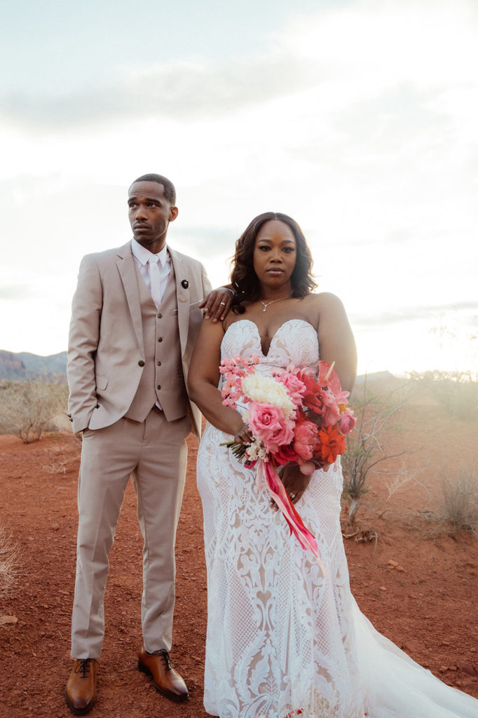 Bold & Colorful in the Vegas Desert. The groom stands behind the bride as he looks out into the desert while the bride holds her bouquet and looks straight 