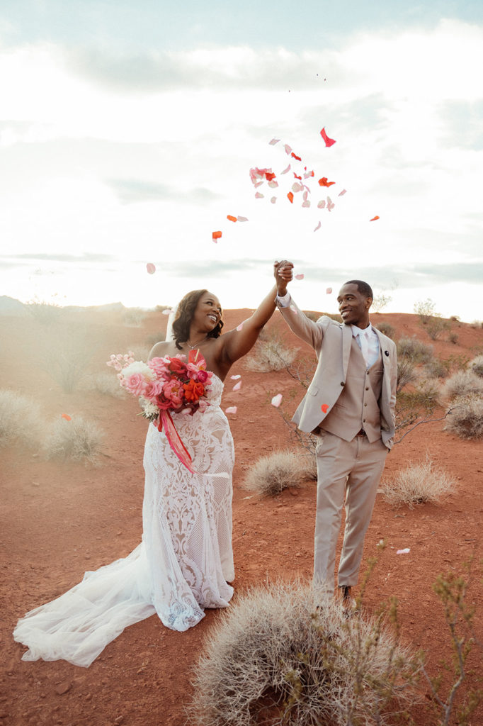 Bold & Colorful in the Vegas Desert. Newlyweds holding hands walking as flower pedals are being thrown in the air 