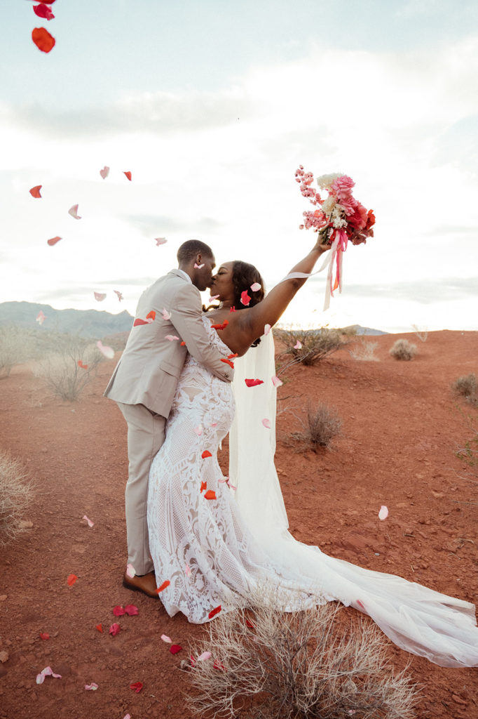 Bold & Colorful in the Vegas Desert. The newlyweds kiss with the bride holding up her bouquet and numerous flower pedals are being thrown in the air around them 