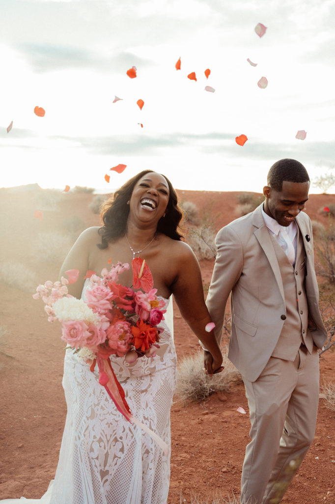 Bold & Colorful in the Vegas Desert. The bride and groom smile and laugh as they walk together holding hands as family and friends throw flower pedals at them 