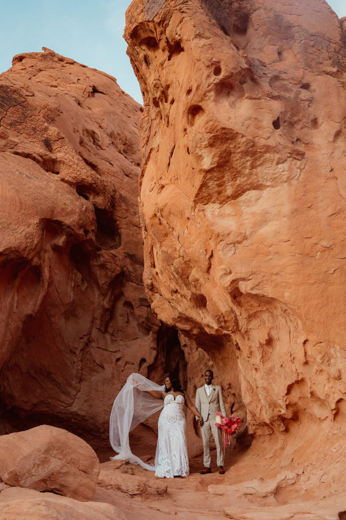 Bold & Colorful in the Vegas Desert. The bride and groom pose for a photo in the middle of a cave in the rocks of valley of fire as the wind blows the brides veil in the wind 
