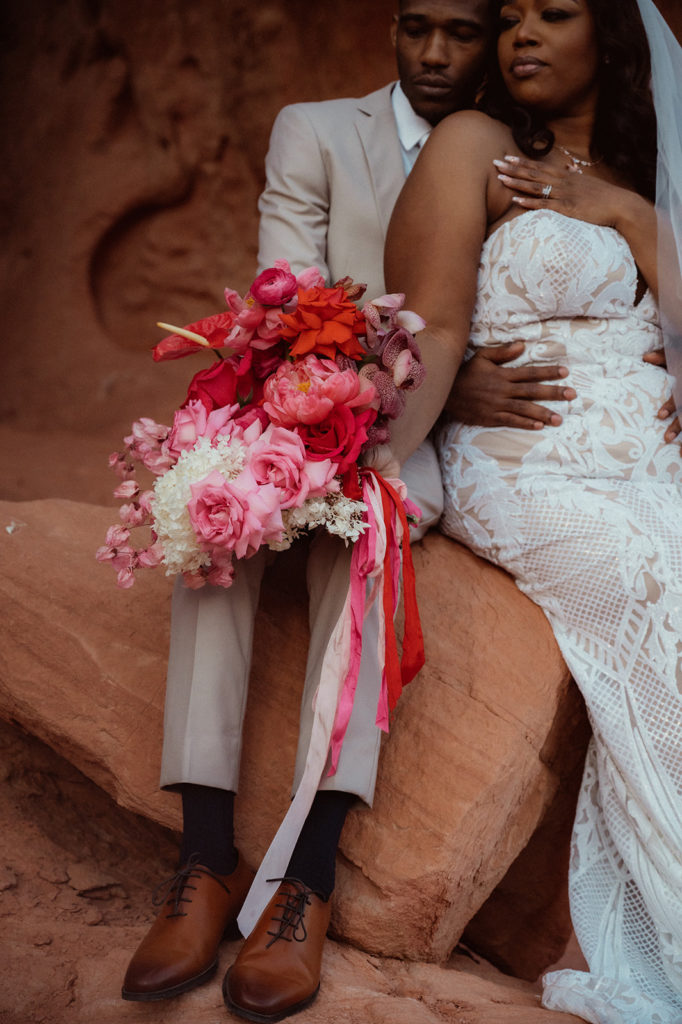 Bold & Colorful in the Vegas Desert. Newlyweds sit on the rock as the bride leans into the groom and they both look off into the distance as she holds her lush monochromatic magenta bouquet 