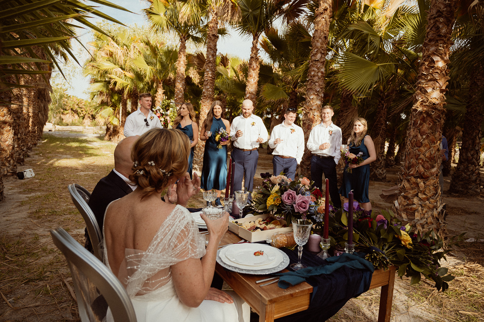 Wedding party doing a cheers with Newlyweds at sweetheart table in palm tree garden 
