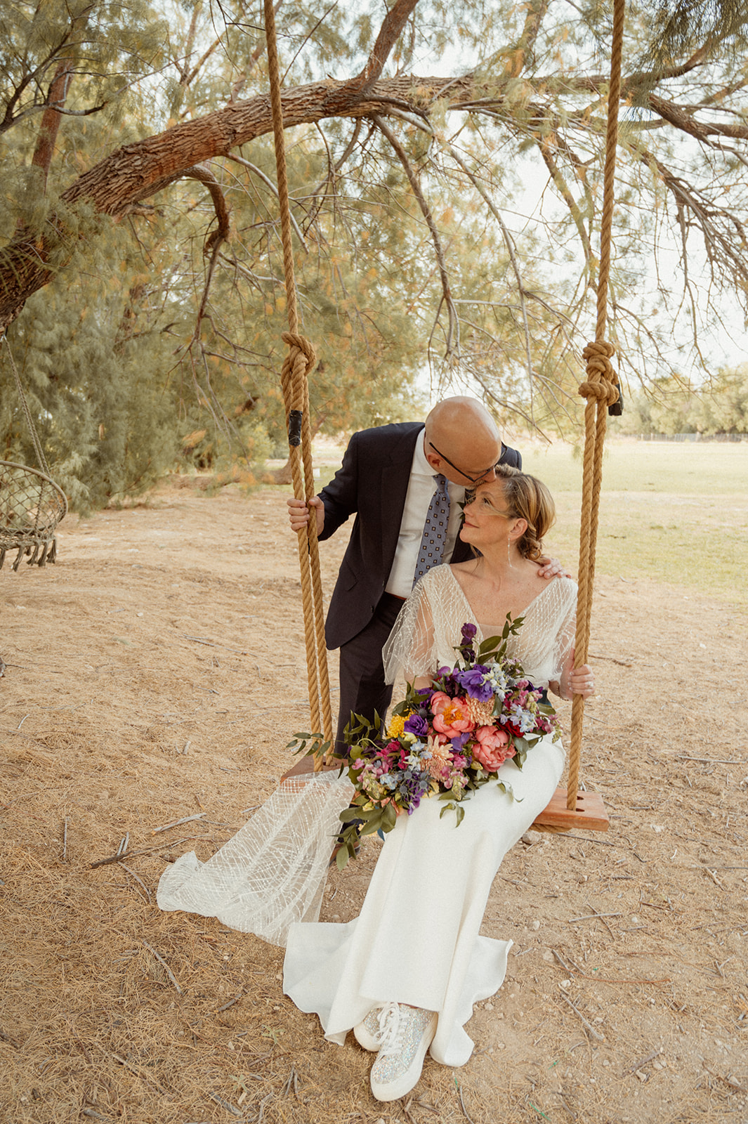 Bride sitting on tree swing while groom kisses her forehead 