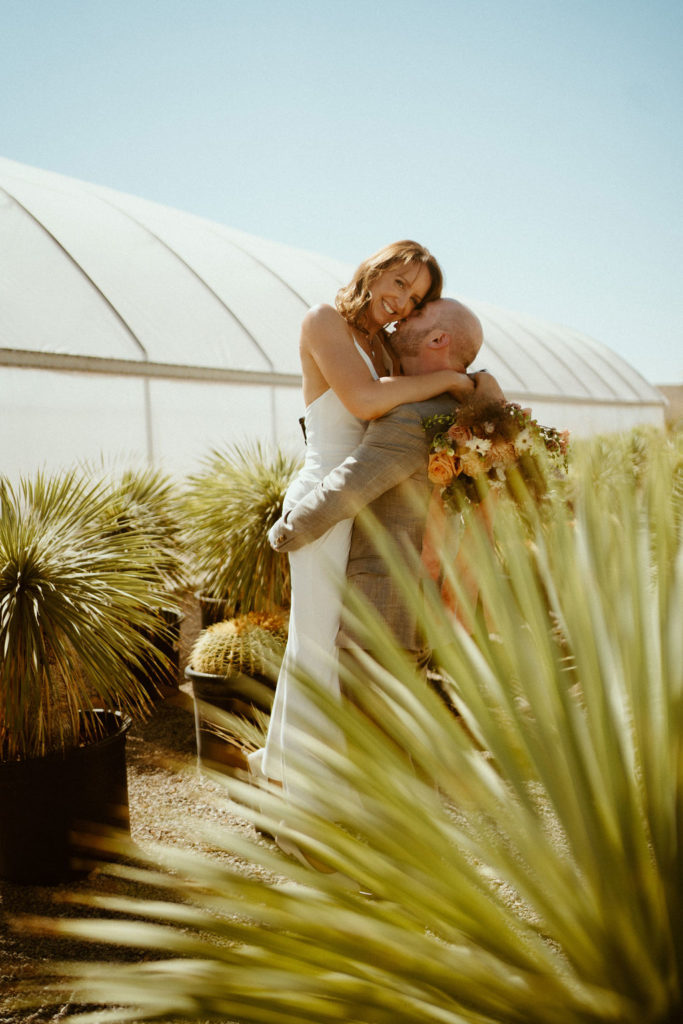 Groom holding up bride, standing next to greenhouse and cactus after getting eloping 