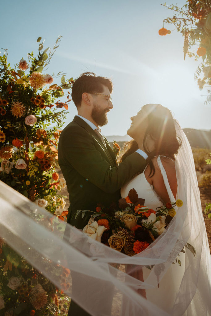 Couple surrounded by veil and fall florals smiling while the sunset is behind them 