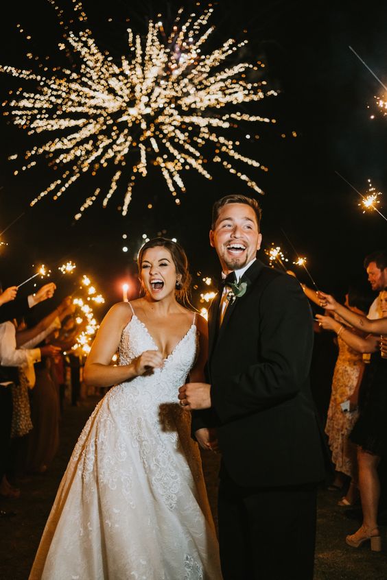 The bride and groom walk down the aisle of guest holding up sparklers as fireworks are going off. 