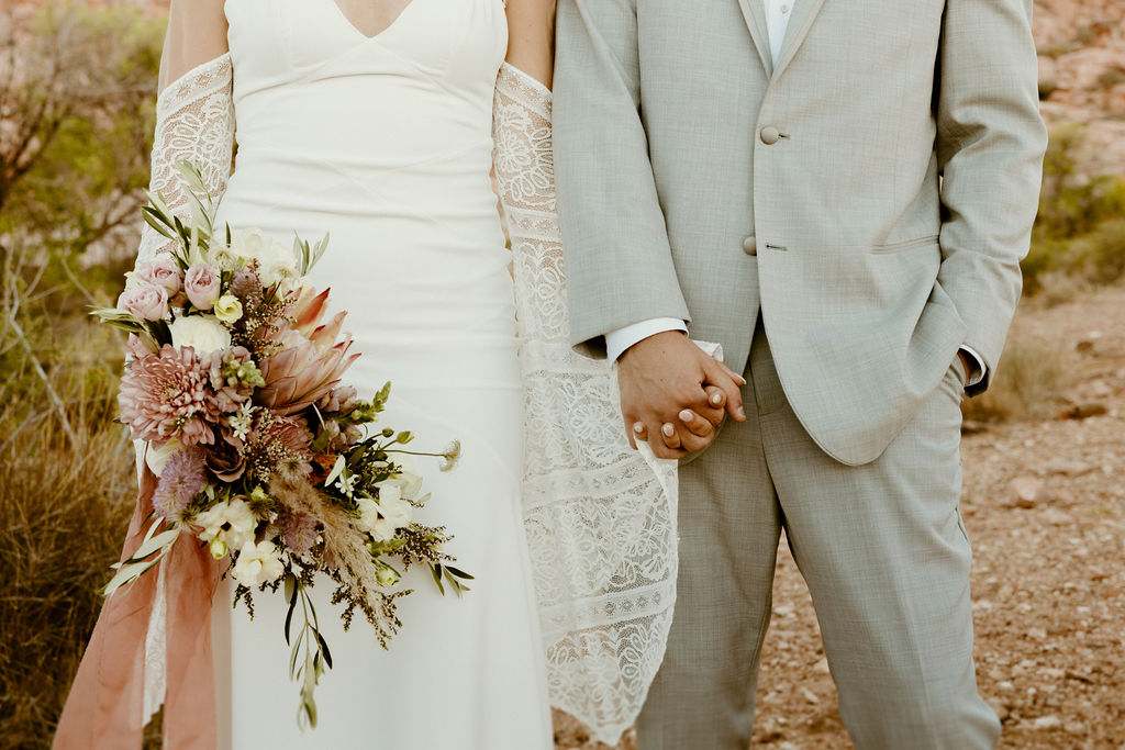 Newlyweds holding hands in the middle of the desert. The bride holding her mauve and light pink bouquet at her side. 