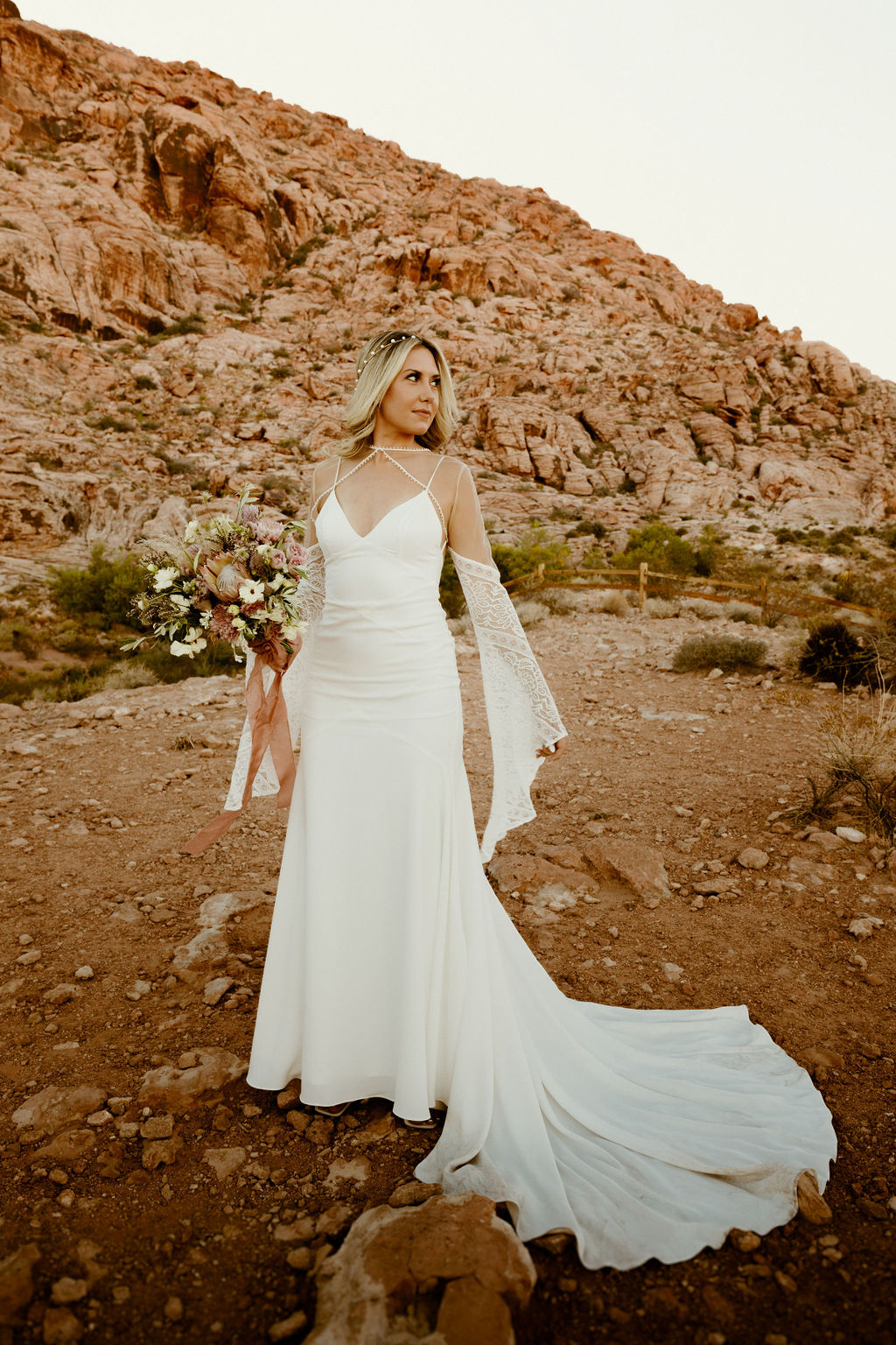 Bride standing in the middle of the desert with her long train and flowing sleeve as she holds her wild flower mauve bouquet 