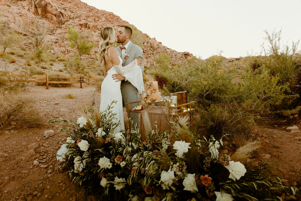 Red Rock Desert & Neon Vegas Lights. Newlyweds kissing next to the cake table in the middle of the desert 