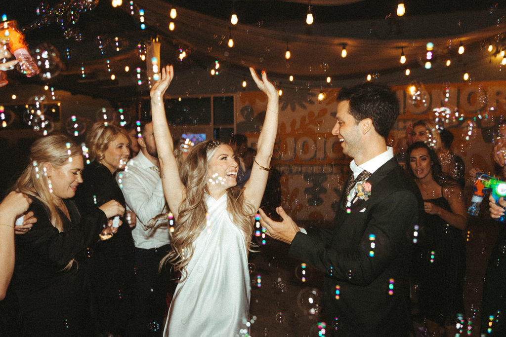 Retro & Colorful Micro-Wedding at the Doyle. Bride and groom cheer as they run through the bubbles in their wedding exit 