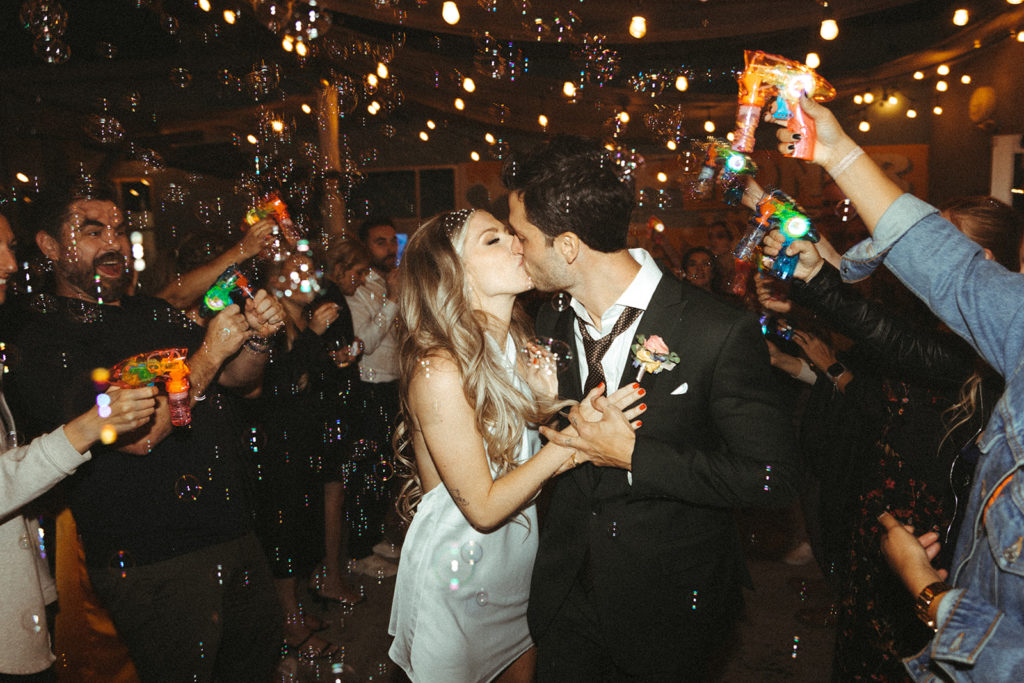 Retro & Colorful Micro-Wedding at the Doyle. Newlyweds stop and kiss in the middle of leaving their reception as friends and family gather around them blowing bubbles