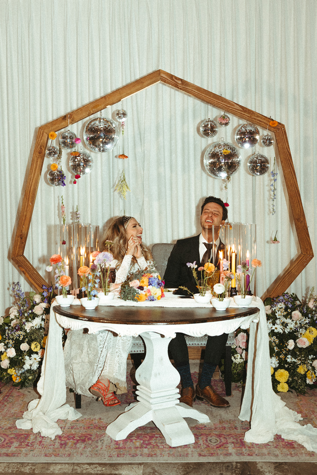 Retro & Colorful Micro-Wedding at the Doyle. Newlyweds sit at the sweetheart table with a hexagon backdrop and hanging disco balls and florals in the arch. 