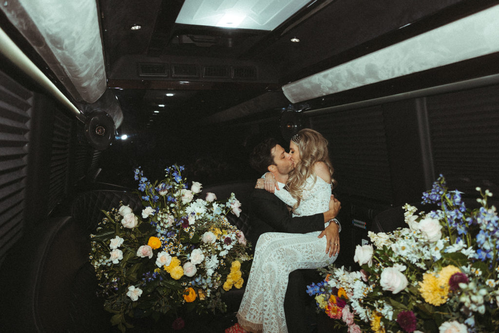 The bride sitting on the grooms lap behind the big color floral arrangements in the back of the party bus kissing 