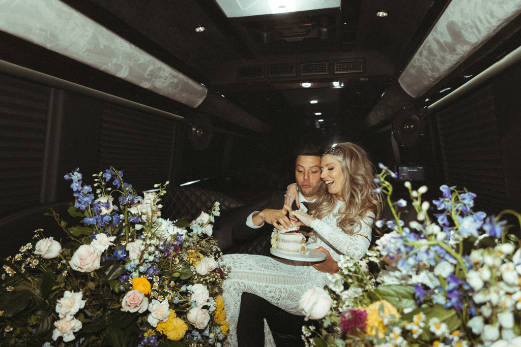 Newlyweds eat cake in the back of the party bus behind all the big and colorful florals  
