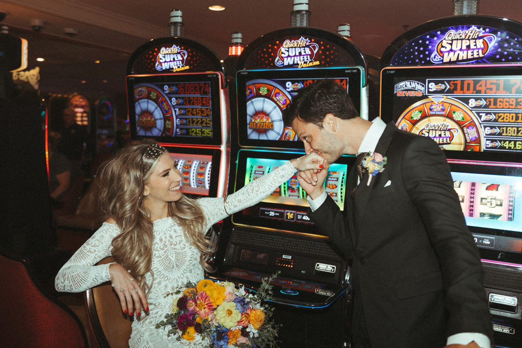 Retro & Colorful Micro-Wedding at the Doyle. Newlyweds hit the Las Vegas casino after their desert oasis ceremony. Groom kisses the brides hand as she sits at the slot machine 