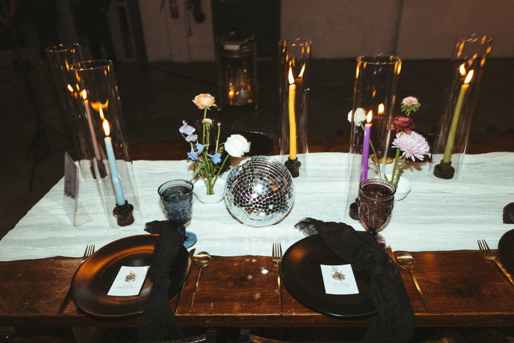 Wooden sweetheart table topped with black plates and bride and groom name cards. Disco balls, colorful taper candles in hurricanes and colorful flowers as the center piece on top of a white cheesecloth runner 
