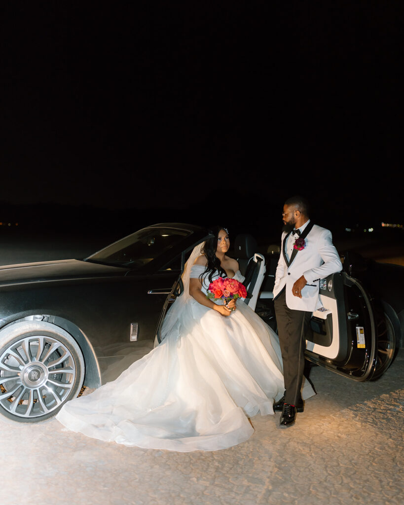 The bride sitting in the Rolls Royce with the door open as the groom leans on the door and looks at his bride 