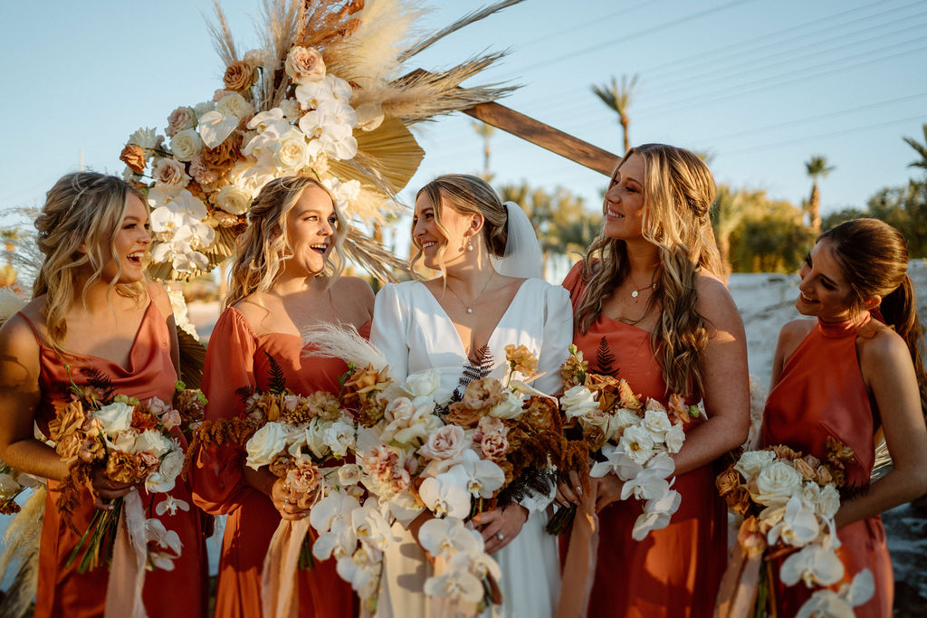 The bride looks at her bridesmaids smiling and laughing. They each hold a hanging boho floral bouquet. 