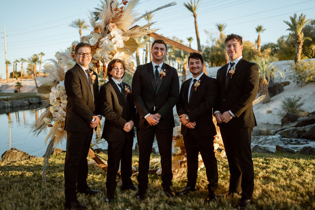 The groom stands in front of the octagon wedding arch with his four grooms men. 
