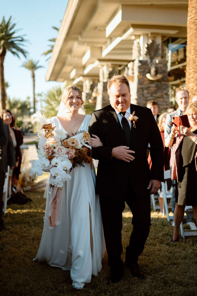 Bride and father walking down the isle as she holds a hanging boho themed bouquet