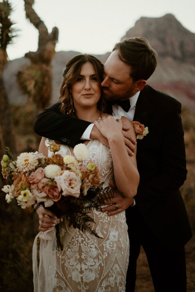 Close up of bride and groom. His arm wrapped around her shoulders. A large cactus in the background. 