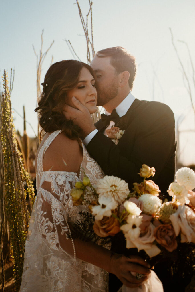 Groom kisses the brides cheek. Her eyes closed holding her bouquet. 