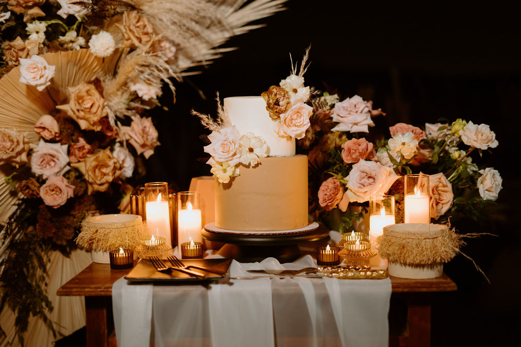 Candles, Pampas, & Romance at Cactus Joe's close up of the wedding cake. Getting specific details of the floral colors. Dusty and soft pink roses. Toffee colored roses and soft white flowers. Giant tan fans and pampas. The table is decorated with white tulle runner and candles. 