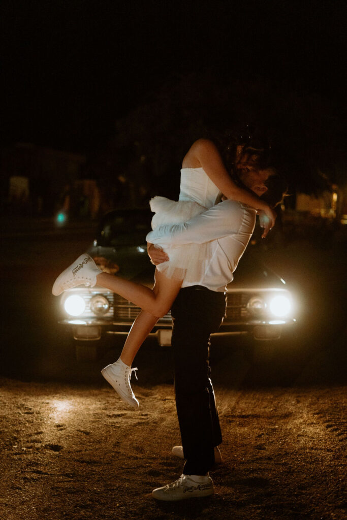 Groom lifting up the bride in her short dress with the headlights of the old school Cadillac shinning on them. 