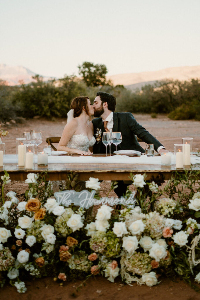 Bride and Groom sitting at the His and Her table sharing a kiss. 