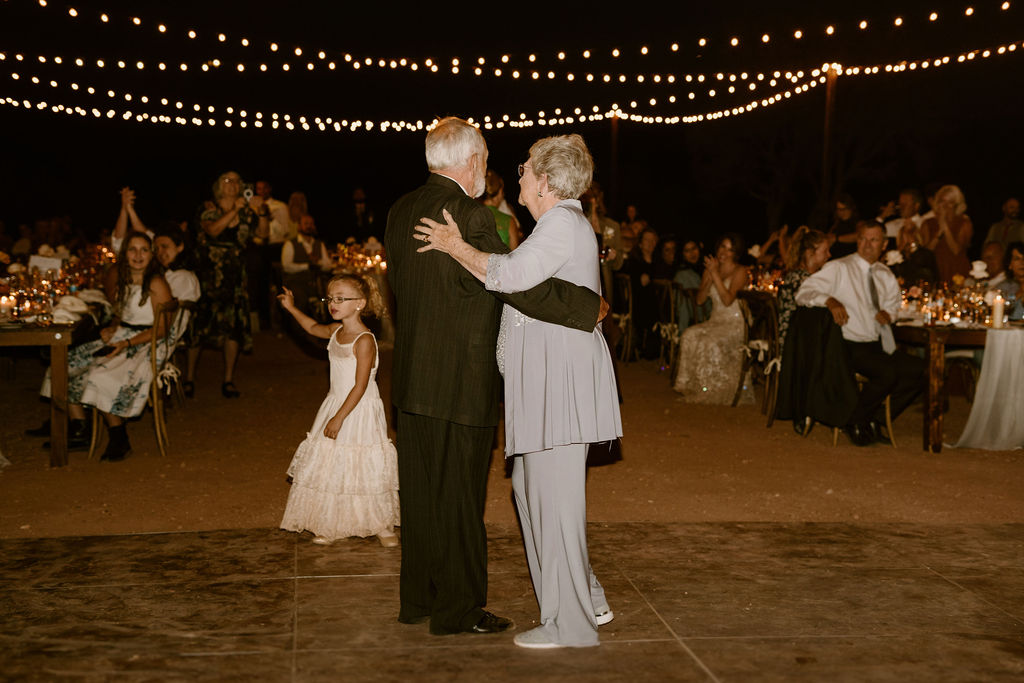 Older couple enjoying a dance at the wedding in the dark with beautiful hanging lights above. A young girl coming in to join them. 