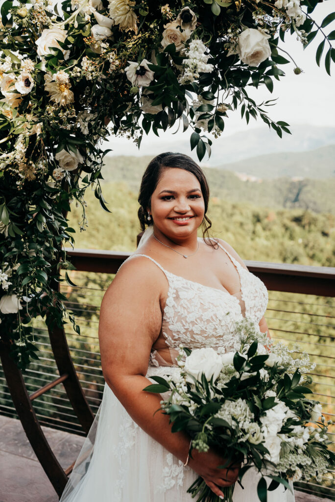 Bride standing in front of the circular arch draped in greenery smiling holding her bouquet 