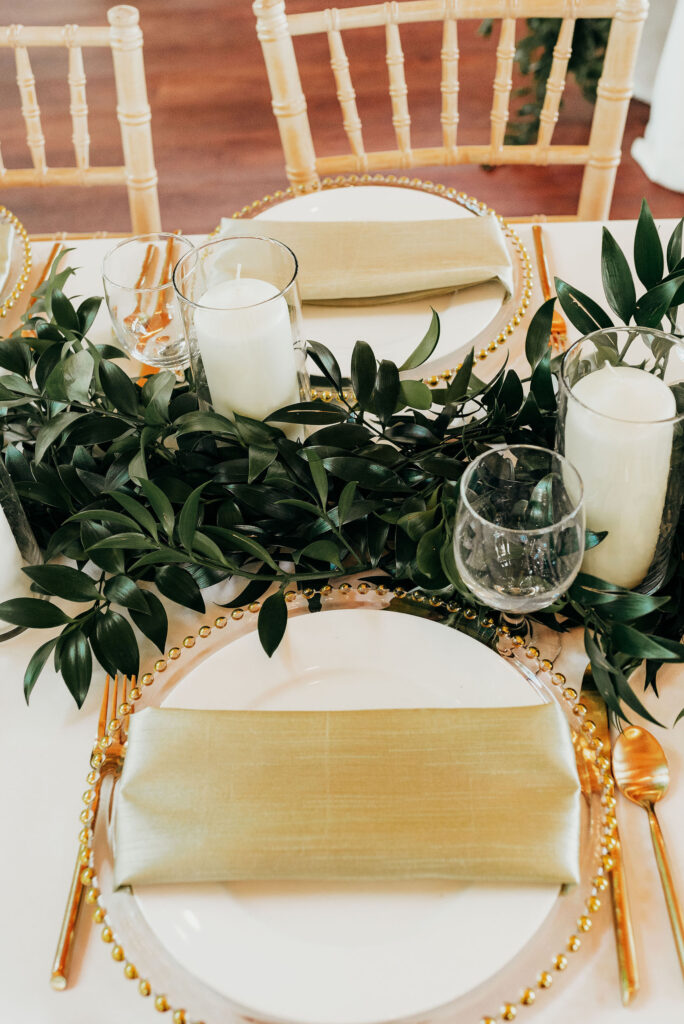 table setting with a clear glass charger and gold trim and a white plate on top and a trifold gold napkin placed in the middle. Gold utensils and candles in hurricanes placed along the greenery runner  