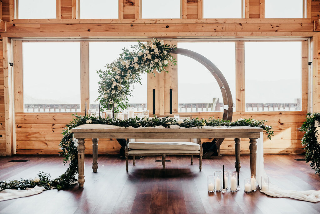 Newlyweds sweetheart table. Wooden circular arch draped with greenery and florals behind. Candles in hurricanes placed all around the sweet heart table with a white sheer runner and a greenery runner placed atop of draped on the side of the table 