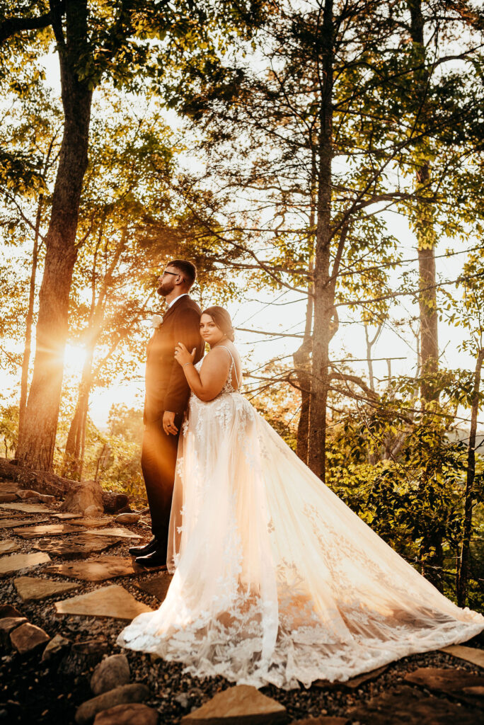 Rolling Hills and Tennessee Whiskey. Bride standing behind the groom holding his arm at the knoll as the sun beams through the trees 