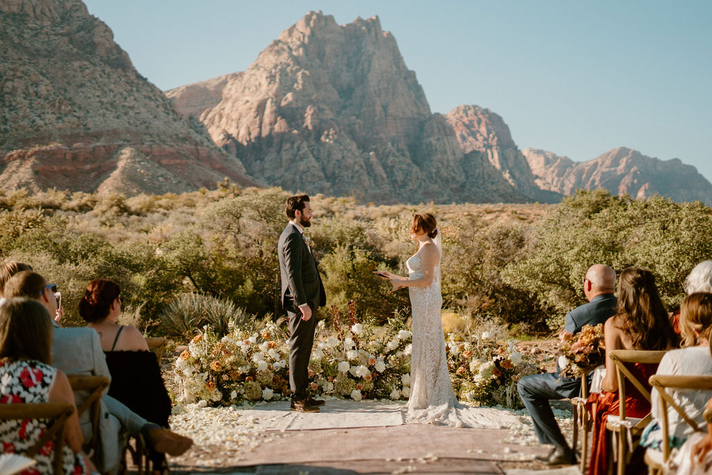 Landscape photo of Bride and Groom reading their vows with the Red Rock Canyon mountains in background and a few guests seen on the side.