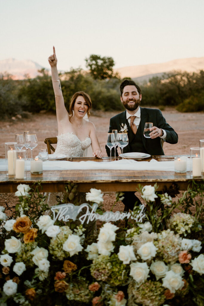 Timeless Red Rock Canyon Wedding Bride and Groom sitting at their his and her table enjoying a drink. Bride is pointing to the sky laughing. 