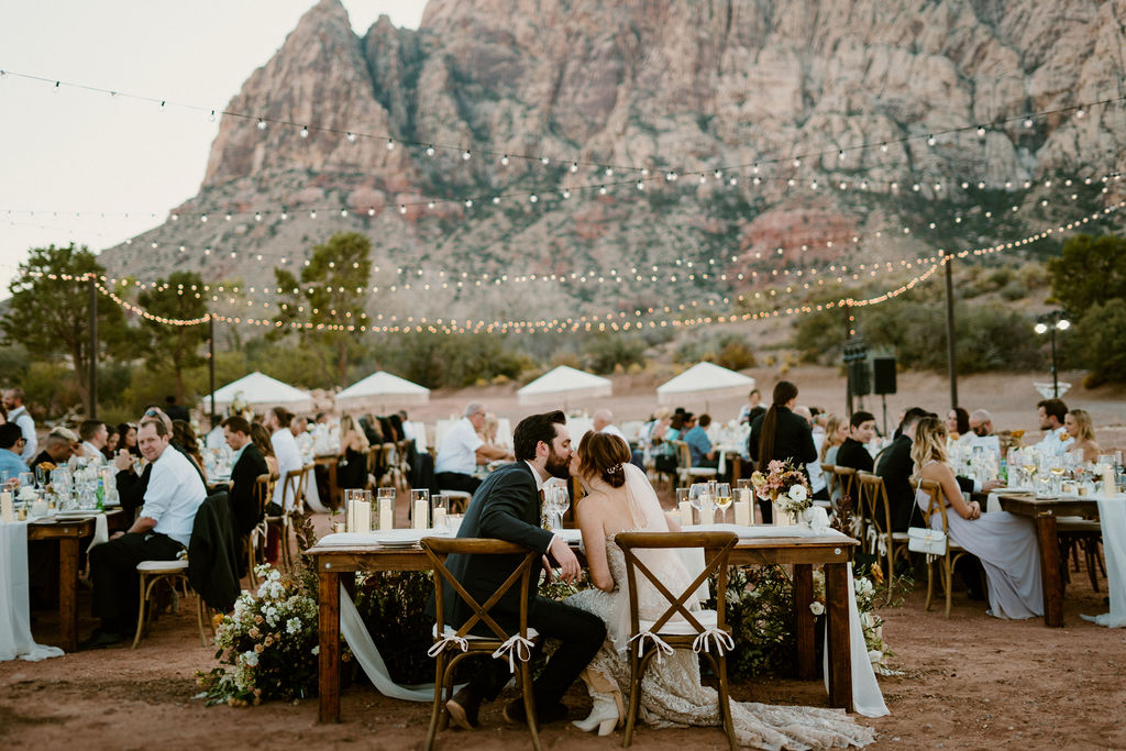 Timeless Red Rock Canyon Wedding looking at the backside of the his and her table while the bride and groom share a kiss. Guests are in the background 